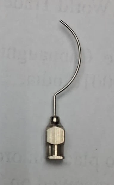 The manufactured cannula laid on a pale ground, seen from the side: a hexagonal Luer hub, a short straight length rising from it, and the long curved portion sweeping away to a fine tip.